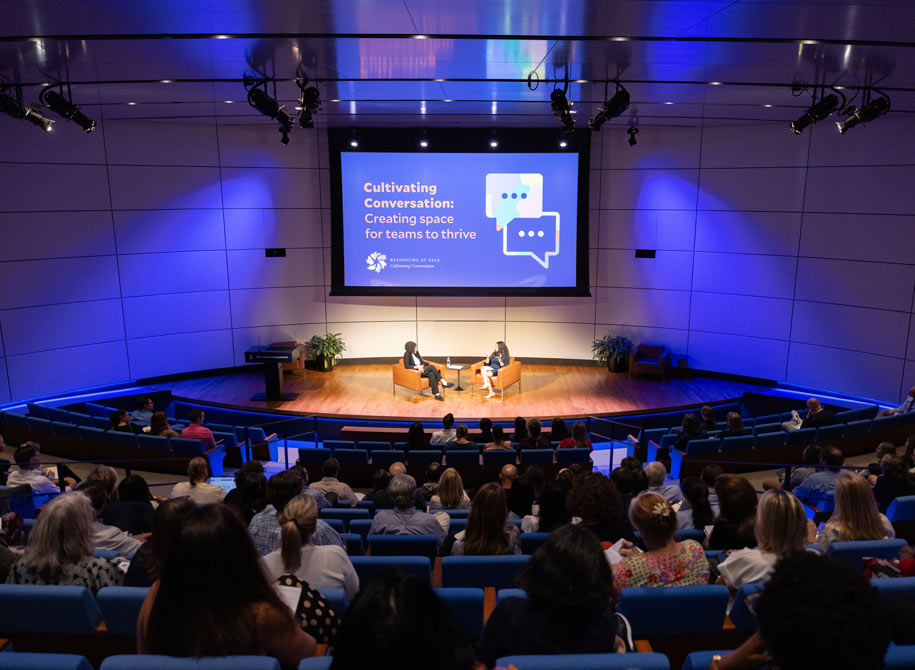 Photo of an auditorium full of Yale Managers with two speakers on stage seated in arm chairs at the cultivating conversation: creating space for teams to thrive