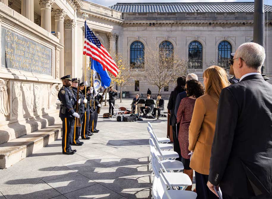 Military or police color guard displaying the american flag in front of the schwarzmen center while the crowd stands and watches and a small band of four plays.