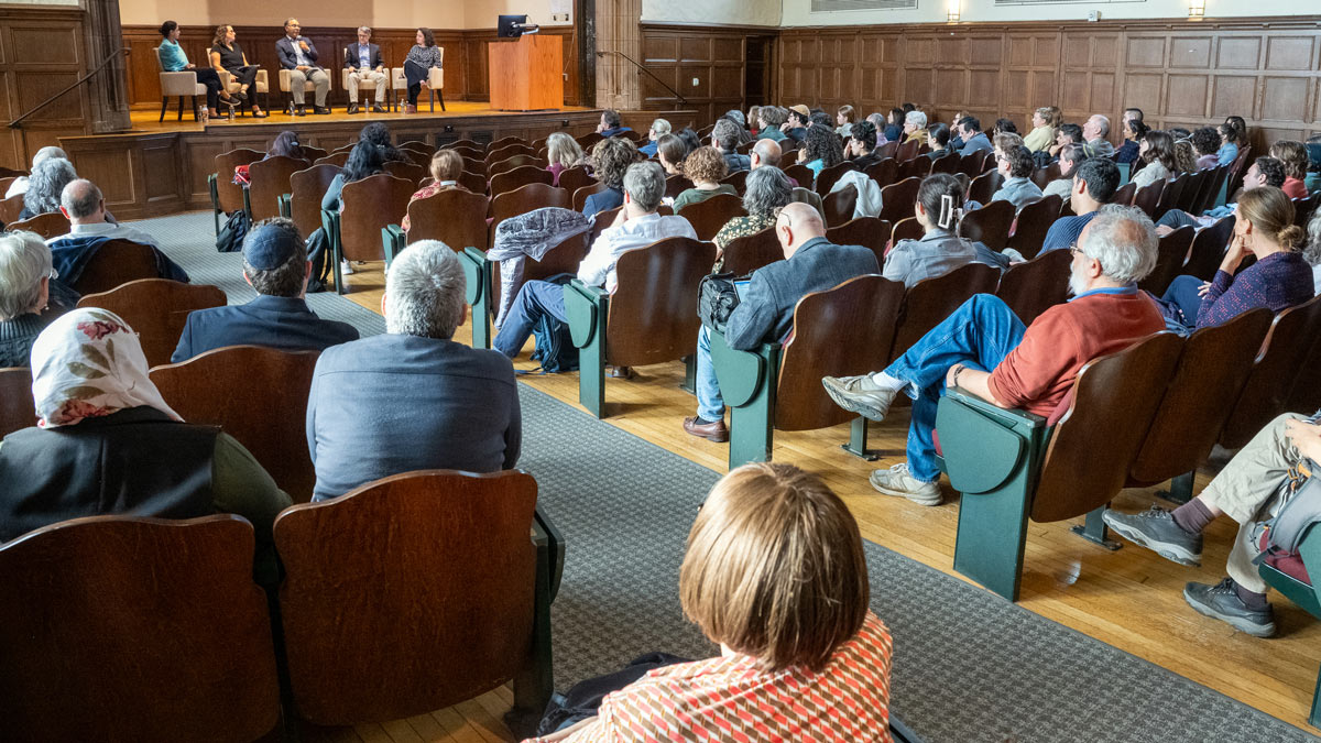 An auditorium filled with seated participants watching a panel of 5 on stage having a discussion.